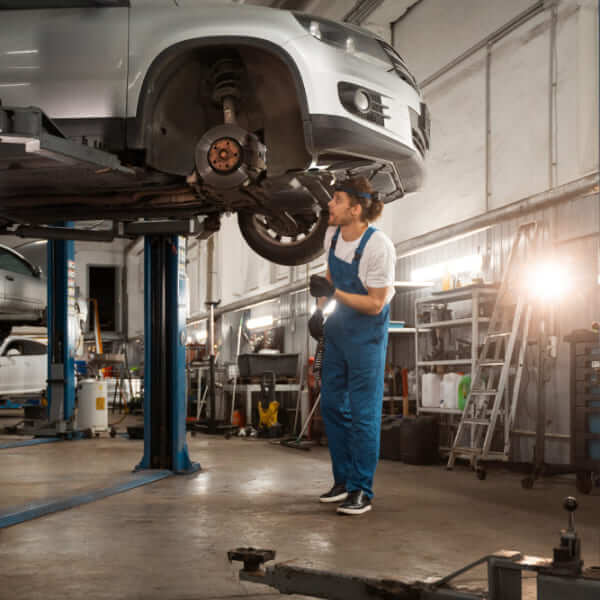 Un mécanicien en bleu de travail examinant le disque de frein et la suspension d'une voiture argentée soulevée par un pont hydraulique dans un garage.