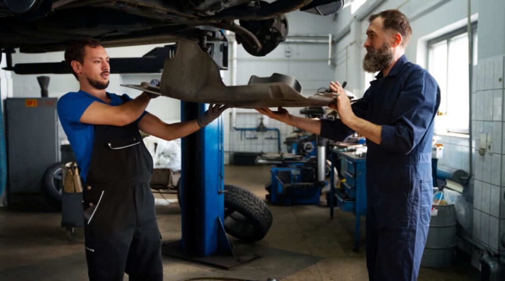 Deux mécaniciens dans un atelier automobile professionnel travaillant ensemble pour retirer une grande protection en plastique du dessous d'une voiture sur un pont élévateur.