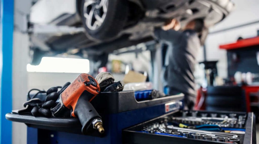 Un gros plan d'un chariot à outils bleu avec une clé à chocs pneumatique orange et divers outils à main, avec un mécanicien flou travaillant sur une voiture en arrière-plan.